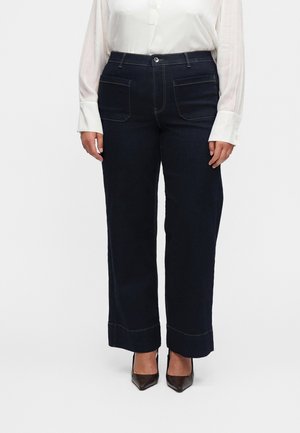 Kvinde iført mørke, brede denimjeans, hvid langærmet skjorte med knapper og sorte spidse højhælede sko, stående mod en ensfarvet baggrund.