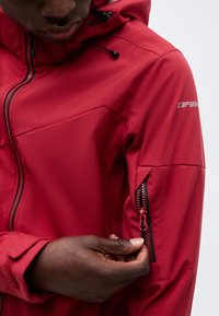 Personne portant une veste rouge à capuche, tenant et fermant une poche zippée sur la manche gauche.