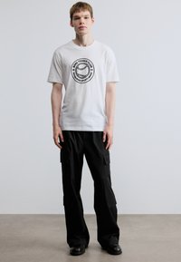 Balti medvilniniai marškinėliai su apvalia grafine teniso kamuoliuko spauda ir užrašu. Derinami su juodomis laisvėjančiomis krovinių kelnėmis su šoniniais kišenėmis.