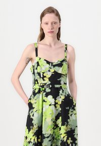 Kukallinen mekko mustalla pohjalla, jossa on abstrakteja vihreitä ja keltaisia kuvioita, muotoiltu rinta, säädettävät olkaimet ja vyötäröllä vyö metalliosalla.