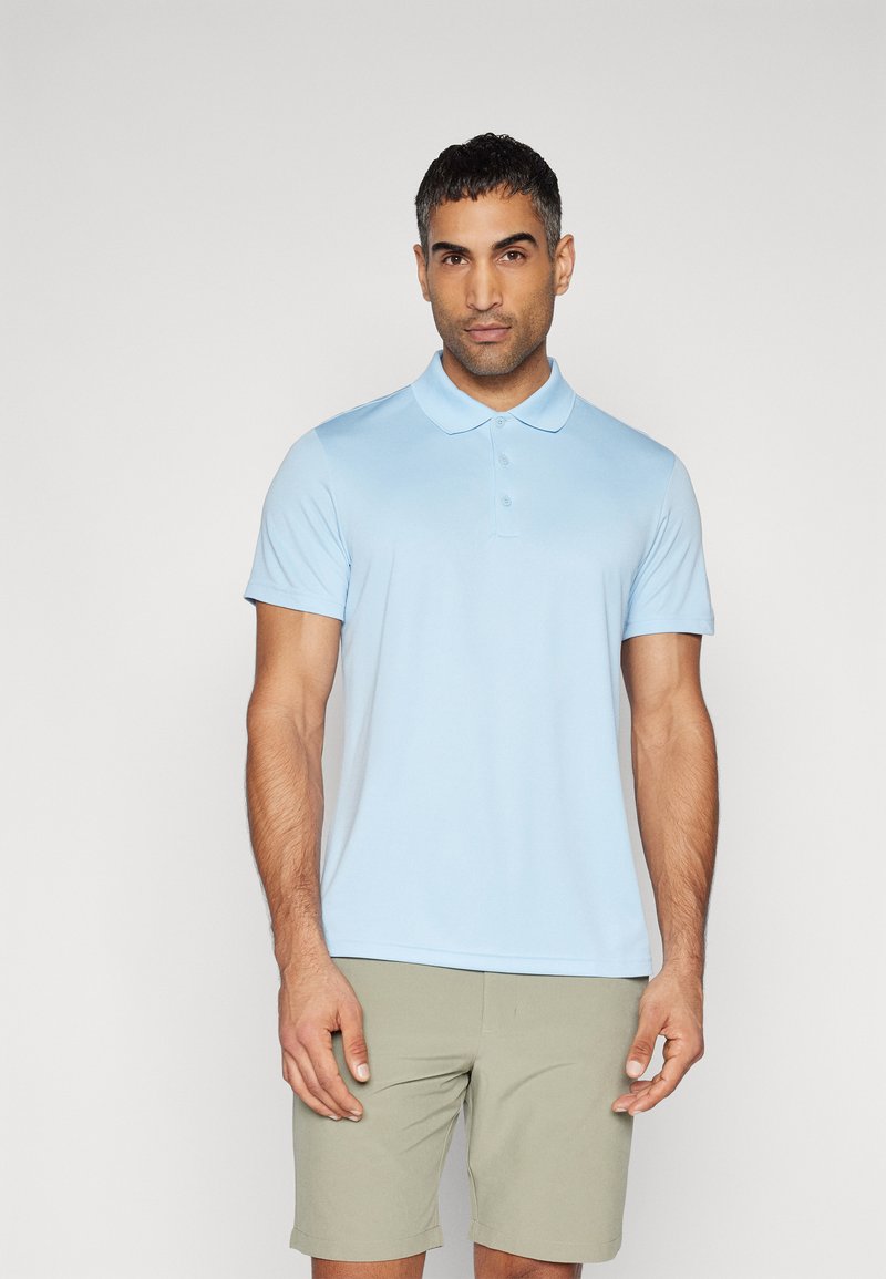Camisa polo azul clara com mangas curtas, colarinho e frente com três botões. Usada com calções verdes claros, de textura suave e corte casual.