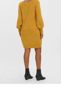 Robe en tricot jaune moutarde à coupe ample, manches bouffantes et poignets côtelés, associée à des bottines noires à talon épais.