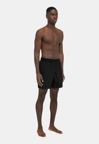 Pantaloni da bagno neri con vita elastica, tasche laterali e vestibilità rilassata, indossati da una persona a piedi nudi.