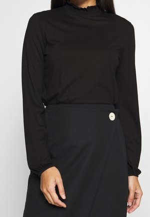 Woman wearing a black long-sleeve top with ruffled collar and cuffs paired with a black wrap skirt secured by a white button.