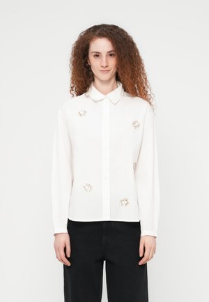 Γυναίκα με κατσαρά κόκκινα μαλλιά, ντυμένη με λευκό πουκάμισο με κουμπιά και κεντήματα σε σχήμα ματιού, και μαύρο παντελόνι, στέκεται μπροστά σε απλό φόντο.