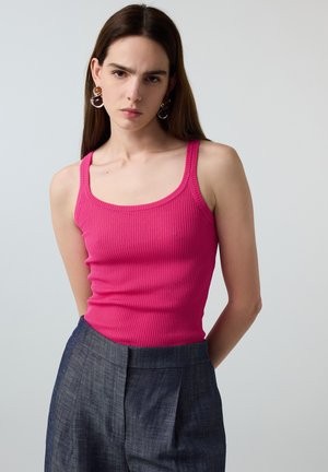 Vrouw met lang bruin haar, grote oorbellen, een felroze geribde tanktop en donkergrijze hoge taille broek, staand tegen een grijze achtergrond.