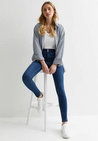 Top blanco de tirantes debajo de una camisa de rayas azul y blanca de gran tamaño, combinada con jeans de tiro alto y zapatillas blancas, sentado en un taburete.