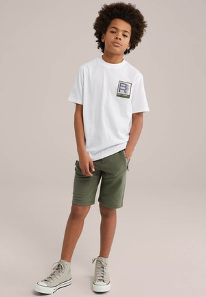 Witte katoenen t-shirt met een logo patch, gecombineerd met groene shorts. Schoeisel met een hoge ontwerp in lichtgrijs met veters. Eenvoudige achtergrond.