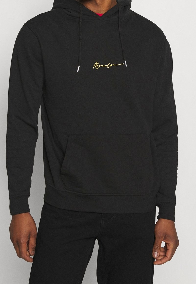 Homme portant un sweat à capuche noir avec un logo jaune en écriture cursive centré sur la poitrine et une poche kangourou à l'avant, assorti à un pantalon noir.