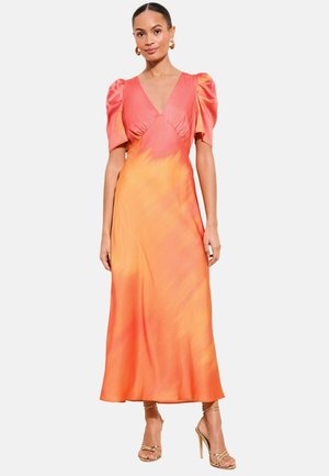 Frau trägt ein Satinkleid in Pink- und Orangetönen mit Puffärmeln und goldenen, hochhackigen Sandalen mit Riemchen, steht vor einem weißen Hintergrund.