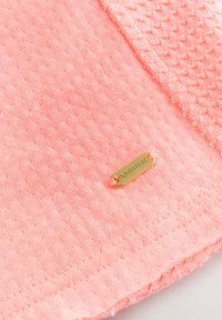 Tissu rose clair avec une finition texturée, comportant une étiquette dorée estampillée "DOWNTIME". Le bord présente un motif décoratif semblable à du crochet.