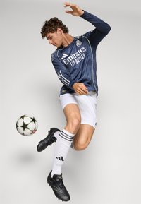 Maglia da calcio a maniche lunghe blu con dettagli gialli, pantaloni corti bianchi, scarpe da calcio nere e calzettoni bianchi a righe, mentre calcia un pallone da calcio.