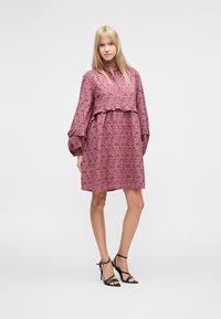 Vestido rosa antigo empoeirado com mangas compridas bufantes, bainha com folhos e padrão floral intrincado. Combinado com sandálias pretas de tiras.