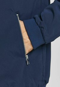 Námořnicky modrá bomber bunda se zipovou kapsou, žebrovanými manžetami a hladkým, texturovaným povrchem látky.