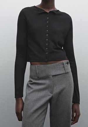 Personne à la peau foncée portant un haut court noir à manches longues et boutonné, ainsi qu’un pantalon gris taille haute avec une ceinture asymétrique.
