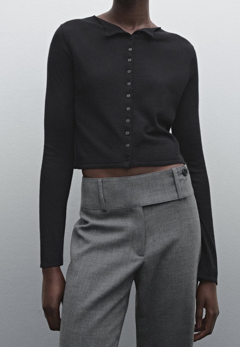 Personne à la peau foncée portant un haut court noir à manches longues et boutonné, ainsi qu’un pantalon gris taille haute avec une ceinture asymétrique.