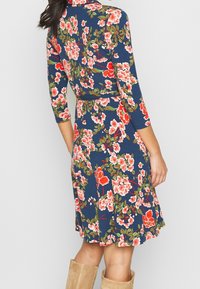 Kvinna i en marinblå knälång klänning med rosa och röda blommönster, trekvartslånga ärmar och sandfärgade mockastövlar, stående mot en plain vägg.