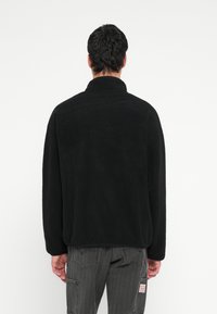 Άνδρας φορώντας μαύρο φλις μπουφάν και ξεθωριασμένο μαύρο παντελόνι cargo, στέκεται με την πλάτη προς το μέρος μας μπροστά σε απλό λευκό φόντο.