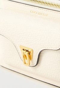 Borsa a mano in pelle con texture cremosa, rifiniture in oro, chiusura a pattina e bordi scuri a contrasto. Presenta un logo impresso sul davanti.