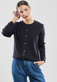 Granatowy dzianinowy kardigan z okrągłym dekoltem, złotymi guzikami z przodu i długimi rękawami. Nosi się go na luźne, dżinsowe spodnie.