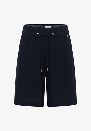 Achteraanzicht van een persoon die zwarte elastische shorts draagt, lichtblauw shirt en beige instapschoenen tegen een witte achtergrond.