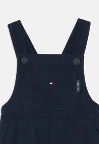 Tengerészkék kordbársony overál széles pántokkal, gombos záródással, elülső zsebbel és egy kis logócímmel, amely piros, fehér és kék színeket tartalmaz.