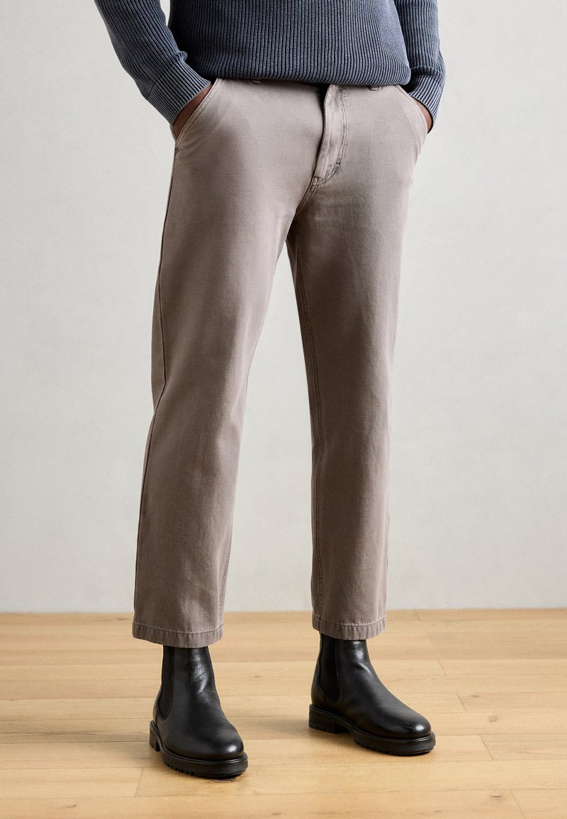 Pantalons en coton beige clair avec une coupe droite, dotés de poches latérales et de détails de couture subtils, associés à des bottines en cuir noires.