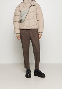 Beige puffjacka med dragkedja och huva, kombinerad med bruna rutiga byxor. Svarta plattformsskor med tjocka soles. Ljust grå axelväska.