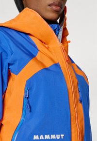 Blaue und orange Jacke mit einem Reißverschluss vorne, kontrastierenden Einsätzen, einer Kapuze und dem Wort "MAMMUT" in Weiß gestickt.