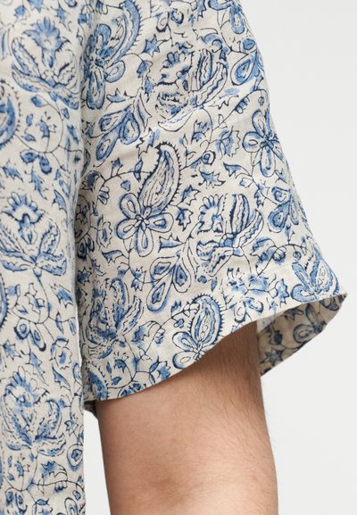 Manche de chemise en tissu léger beige avec motif cachemire bleu. Coupe courte et effilée avec ourlet roulé. Texture douce, accents floraux détaillés.