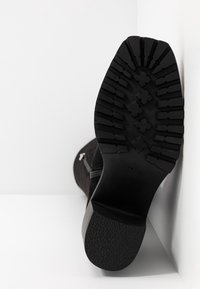 Semelle de botte noire avec motifs de crampons profonds et talon bloc, botte positionnée verticalement contre un mur d'angle blanc et le sol.