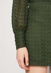 Grön, texturerad klänning av spets med ett geometriskt mönster och långa puffärmar. Ärmsluten och nederkanten är avslutade med en ren kant.