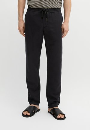 Mężczyzna ubrany w czarne spodnie ze sznurkiem, czarne klapki typu slide oraz czarno-białą koszulę w paski, stojący na jasnym, jednolitym tle.