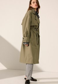 Trench coat verde oliva con vita cin cin, ampi rever e polsini abbottonati; indossato sopra pantaloni grigi e mocassini neri. Texture liscia.