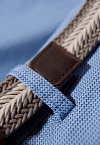 Vävd bälte med ett ljust blått tyg med ett strukturerat mönster, kompletterat med flätade ljus- och mörkbruna accenter samt en mörkbrun läderlapp.