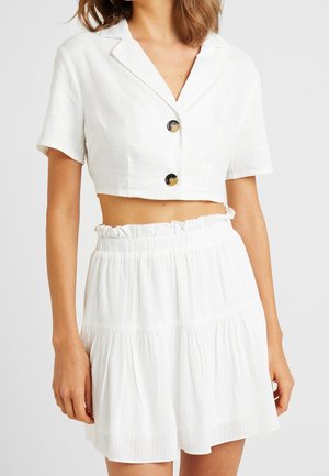 Femme portant une chemise blanche courte à manches courtes boutonnée et une mini-jupe assortie taille haute froncée.