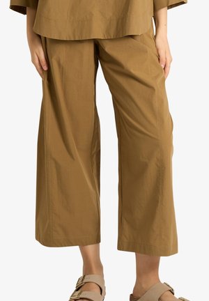 Persona che indossa pantaloni marroni larghi e corti con sandali beige con fibbia, in piedi con le mani rilassate lungo i fianchi su uno sfondo bianco semplice.