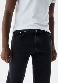 Jeans de mezclilla negra con un diseño clásico de cinco bolsillos, con herrajes plateados y una textura ligeramente deslavada. Llevados con una camiseta blanca lisa.