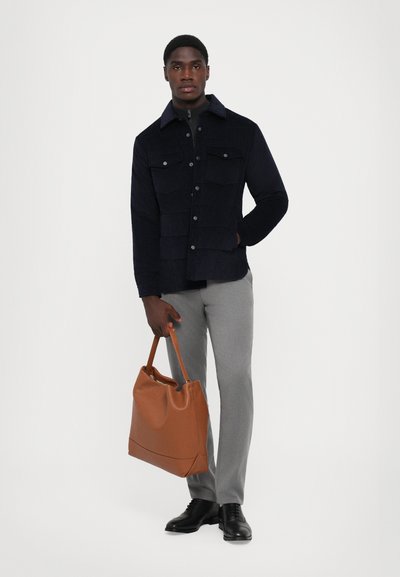 Marineblaues Cord-Jackett mit aufgesetzten Taschen, graue Hose, schwarze Schuhe und braune Tragetasche. Die Stofftextur und der Farbkontrast sind bemerkenswert.