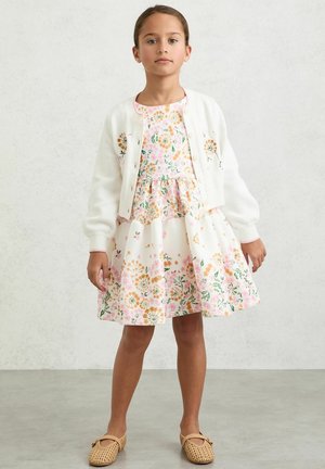 Junges Mädchen trägt ein weißes Blumenmusterkleid, eine weiße, mit Blumen bestickte Strickjacke mit Knöpfen und beigefarbene gewebte Ballerinas, steht auf grauem Boden.