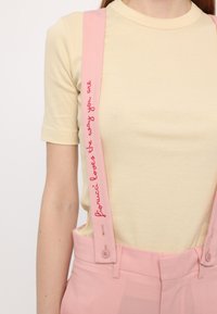 Pink suspenders with embroidered text, attached to a light yellow ribbed t-shirt, paired with light pink trousers featuring a button and pocket.