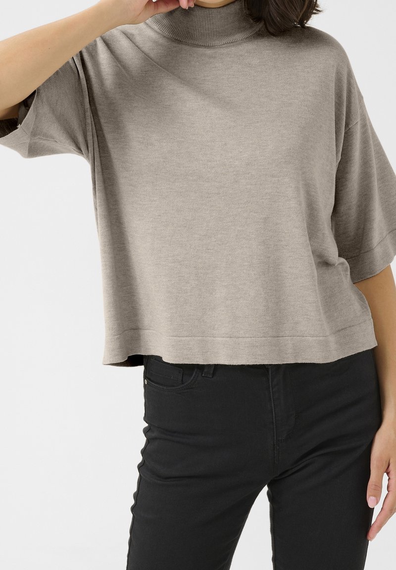 Personne portant un pull à col roulé beige ample à manches courtes et un jean noir ajusté, posant avec un bras levé près du visage.