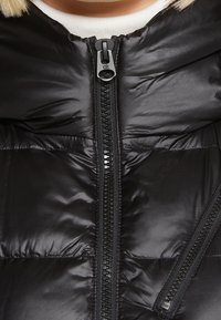 Sort dunvest med en fyldig tekstur, der har en lynlås i fuld længde, mærket glider og en hætte. Materialet virker glat og skinnende.