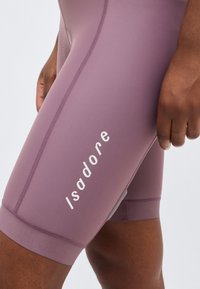 Κοντινό ενός ατόμου που φοράει μοβ σορτς ποδηλασίας με λευκό λογότυπο "Isadore" κατά μήκος του μηρού, το χέρι ακουμπά χαλαρά στο πλάι.