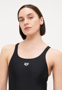 Maillot de bain noir en matériau lisse et extensible. Il présente un décolleté arrondi, des fines bretelles et un logo blanc sur la poitrine. Design simple.
