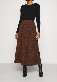 Vrouw in een zwarte lange mouwen top, een bruin en zwart gemarmerde midi rok, en zwarte enkel laarzen staand tegen een effen achtergrond.