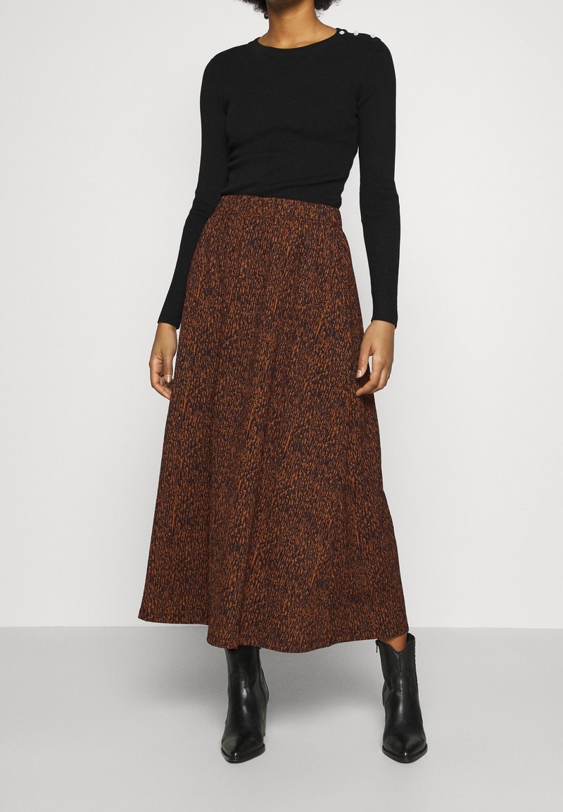 Vrouw in een zwarte lange mouwen top, een bruin en zwart gemarmerde midi rok, en zwarte enkel laarzen staand tegen een effen achtergrond.