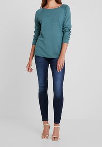 Top de manga larga color verde azulado con un cuello redondo, combinado con jeans ajustados de color azul oscuro y sandalias de tiras beige. Textura suave y diseño ajustado.