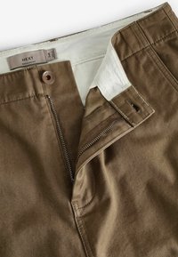 Bruine katoenbroek met een ritssluiting en knoopsluiting, voorzien van een zichtbare binnenste witte voering en gedetailleerde stiksels langs de naden.