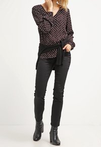 Blouse noire à motifs avec un décolleté en V et des manches longues, associée à un pantalon ajusté noir et des bottines. Présente un détail de ceinture nouée.